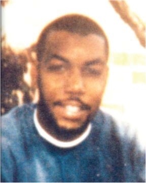 Close-up of a man with dark hair and a beard, wearing a blue shirt, smiling slightly at the camera.