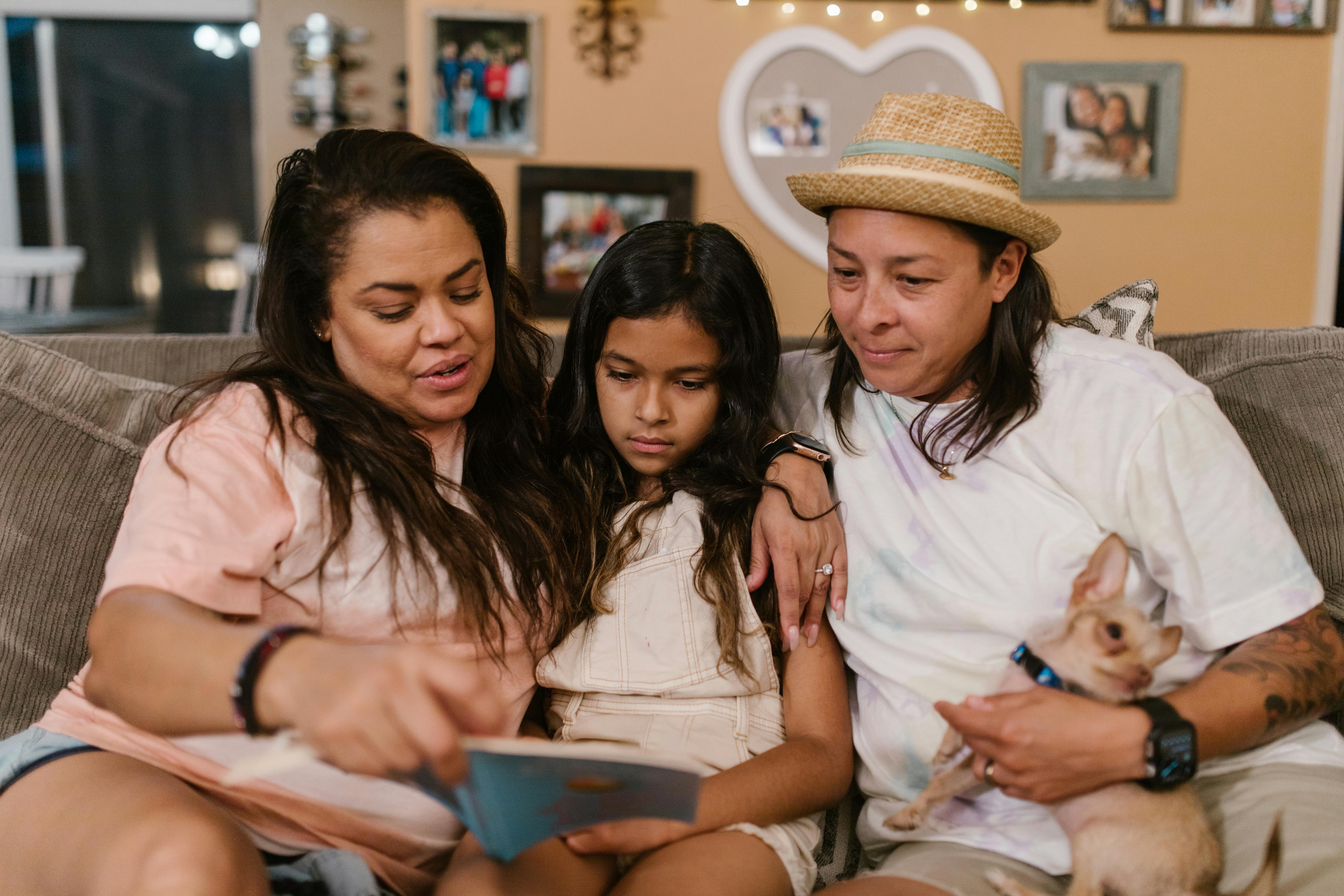 Two mothers and their child read a book together on the couch, with a small dog in their arms.