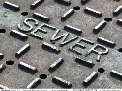 Close-up of a sewer cover with raised lettering spelling 'SEWER' and a textured, patterned surface.
