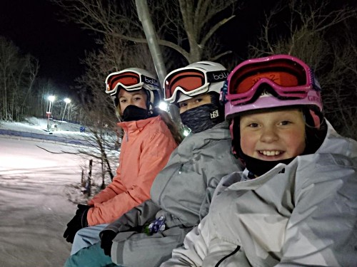Three children wearing ski gear on a chairlift at night, smiling at the camera.