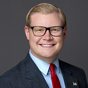 Headshot of a man with blonde hair wearing glasses, a suit, and a red tie against a gray background.
