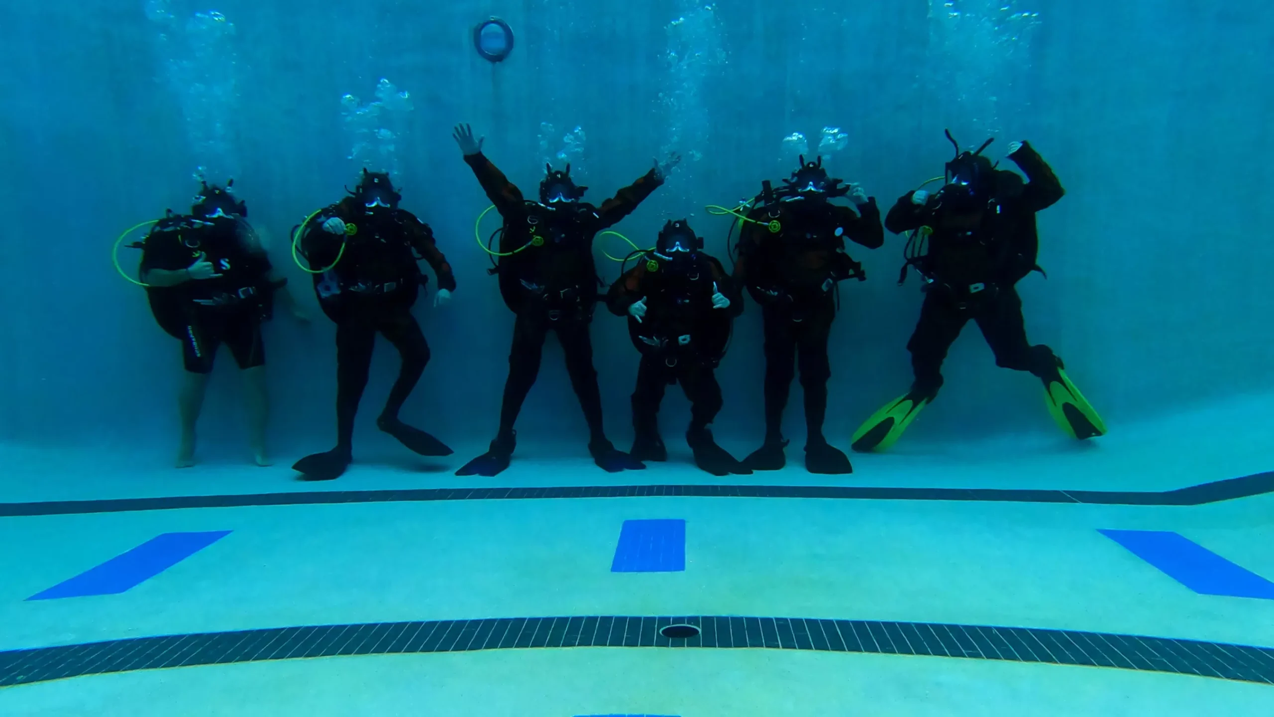 Group of six scuba divers in a pool, posing underwater with scuba gear and bubbles rising.