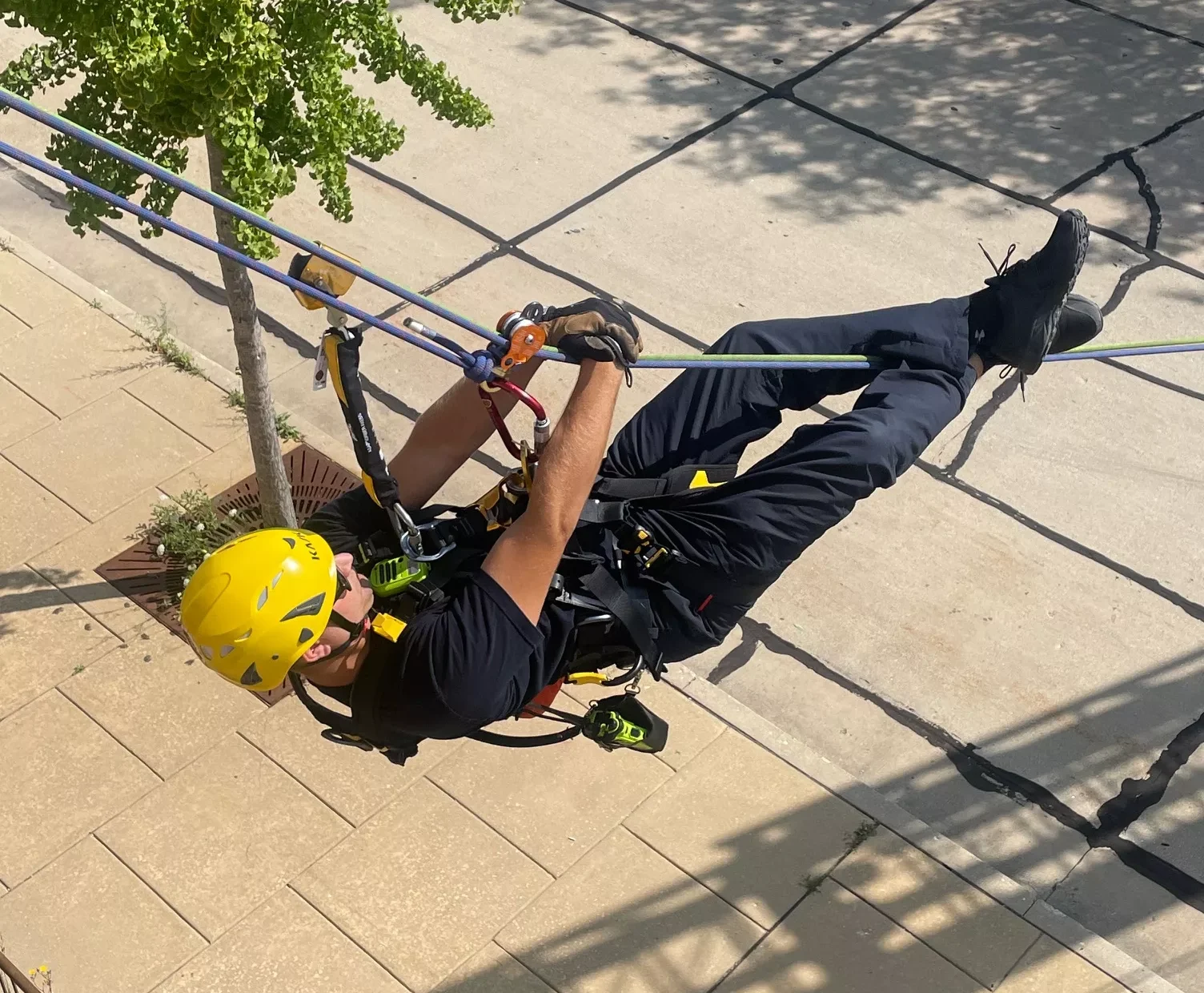 Man in safety harness suspended by ropes, demonstrating rope access techniques, wearing a yellow helmet.