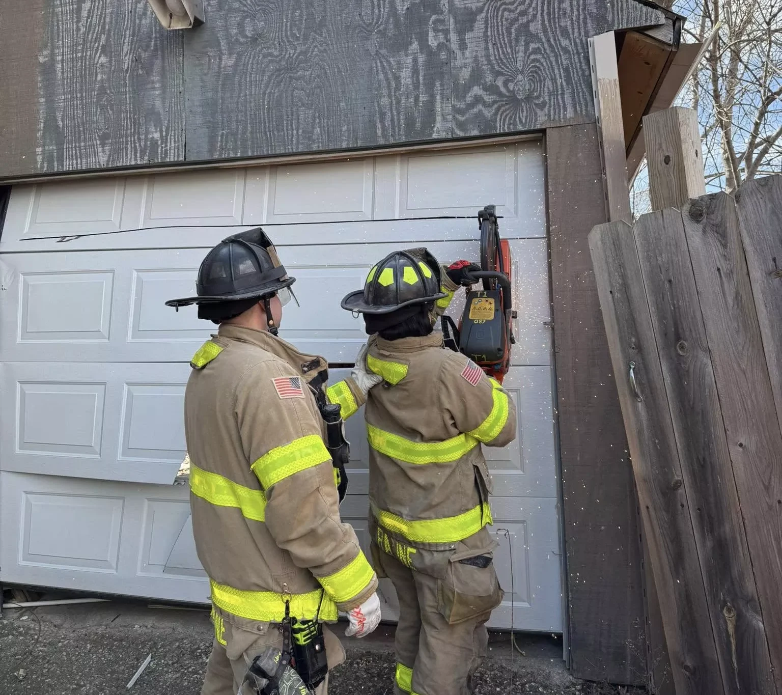 Two firefighters using a saw to cut open a damaged white garage door during an emergency response.