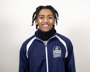 Portrait of a young man with dreadlocks wearing a navy jacket with a community safety logo, smiling against a white