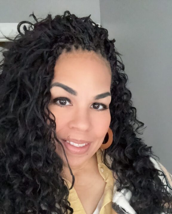 Woman with long, dark, curly hair and brown hoop earrings smiling at the camera. She is wearing a yellow shirt.