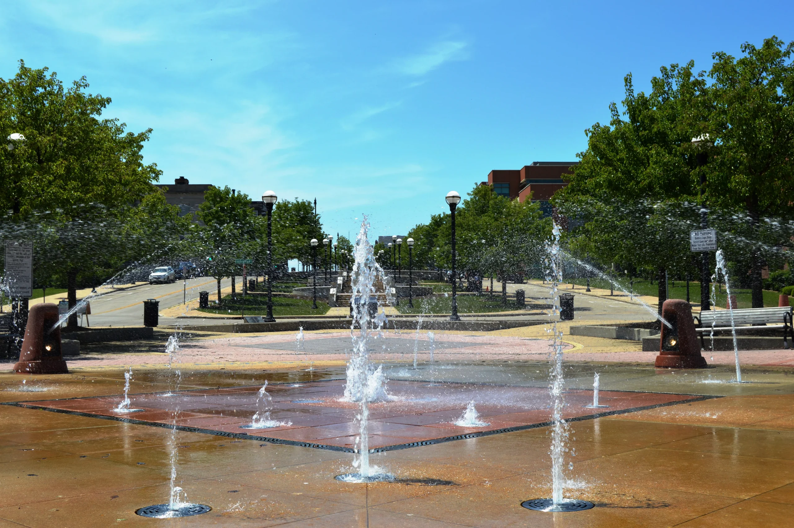 Interactive splash fountain in a public park with trees and buildings in the background on a sunny day.