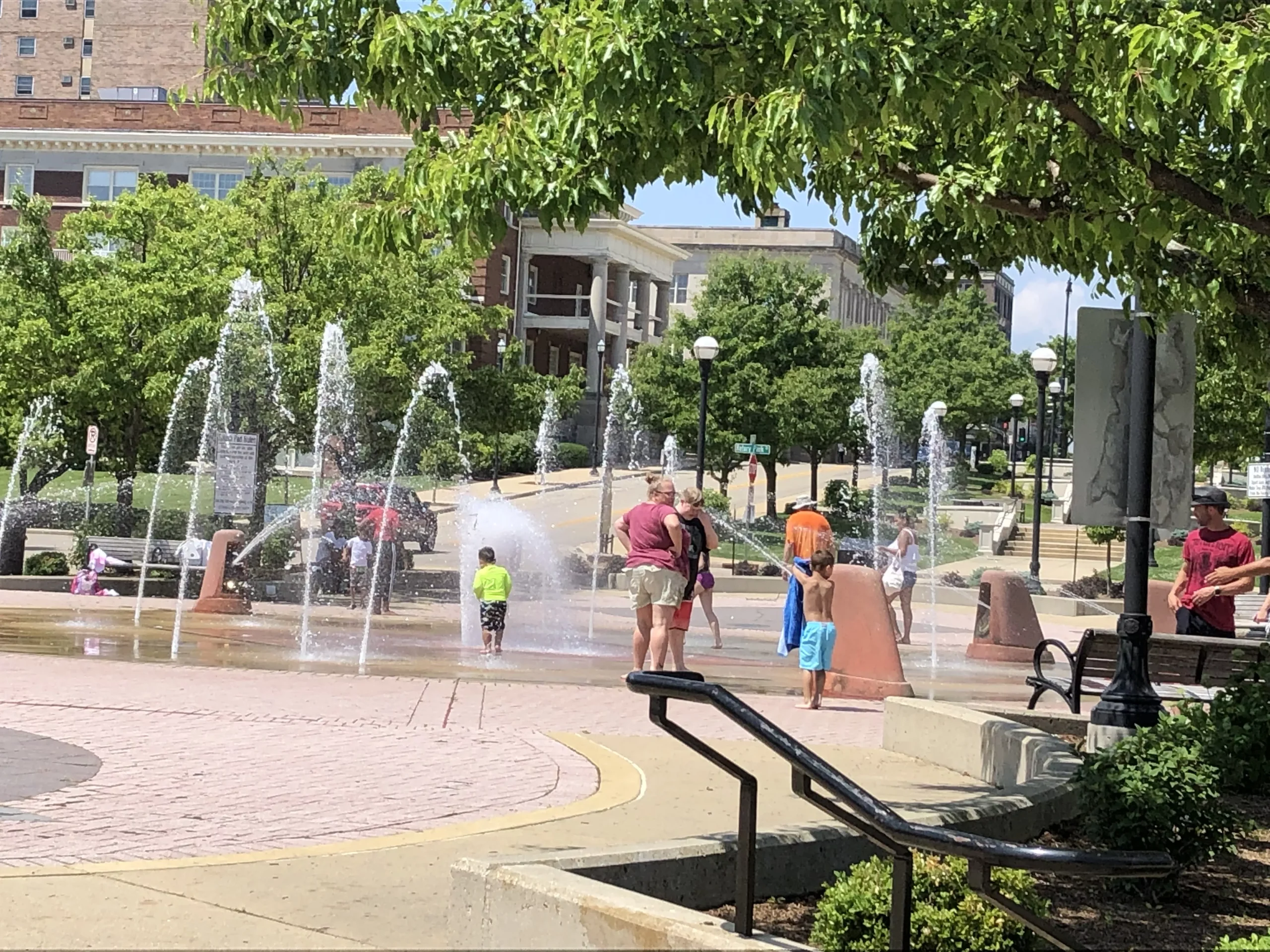Children and adults playing in a public splash fountain on a sunny day in a city park.
