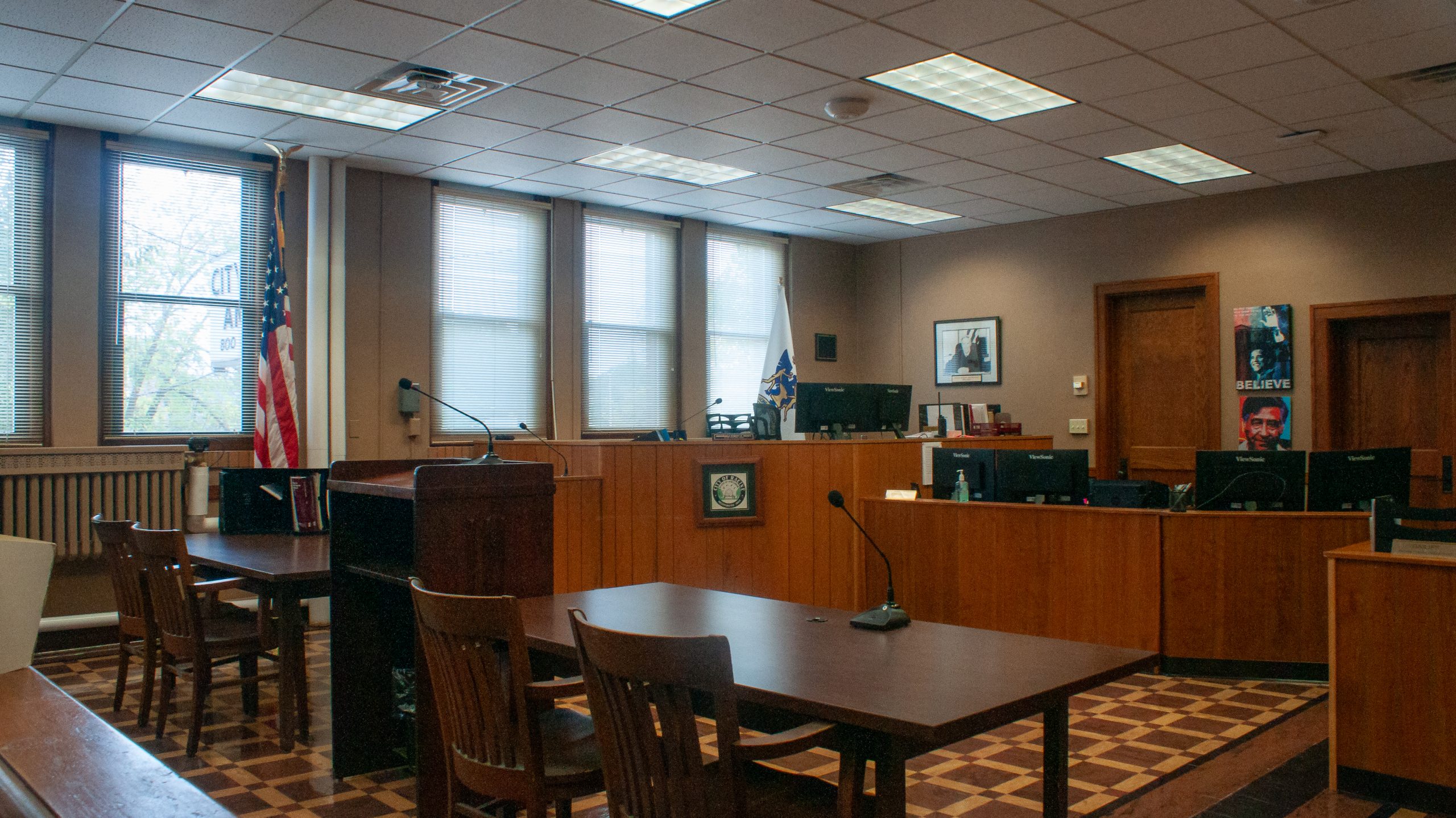 Interior view of a courtroom with tables, chairs, and the American flag visible through the window.