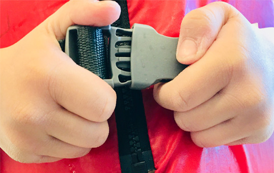 Hands fastening the gray buckle of a red life jacket. Close-up view of the buckle being secured.