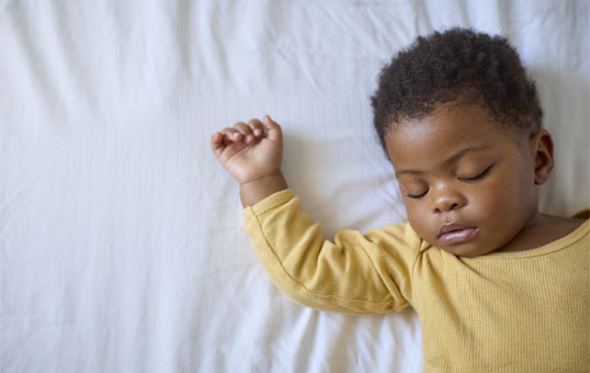 Infant sleeping peacefully on white bedding, wearing a yellow shirt. Overhead shot with copy space.