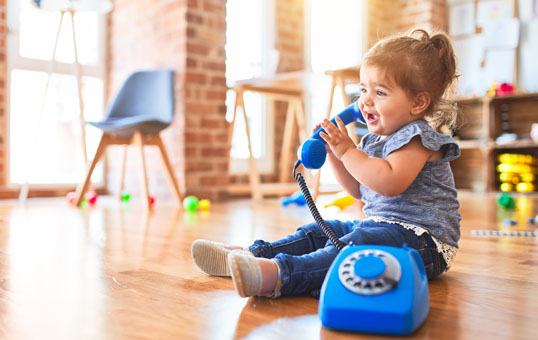 Toddler girl joyfully plays with a blue toy telephone while sitting on a wooden floor in a bright room.