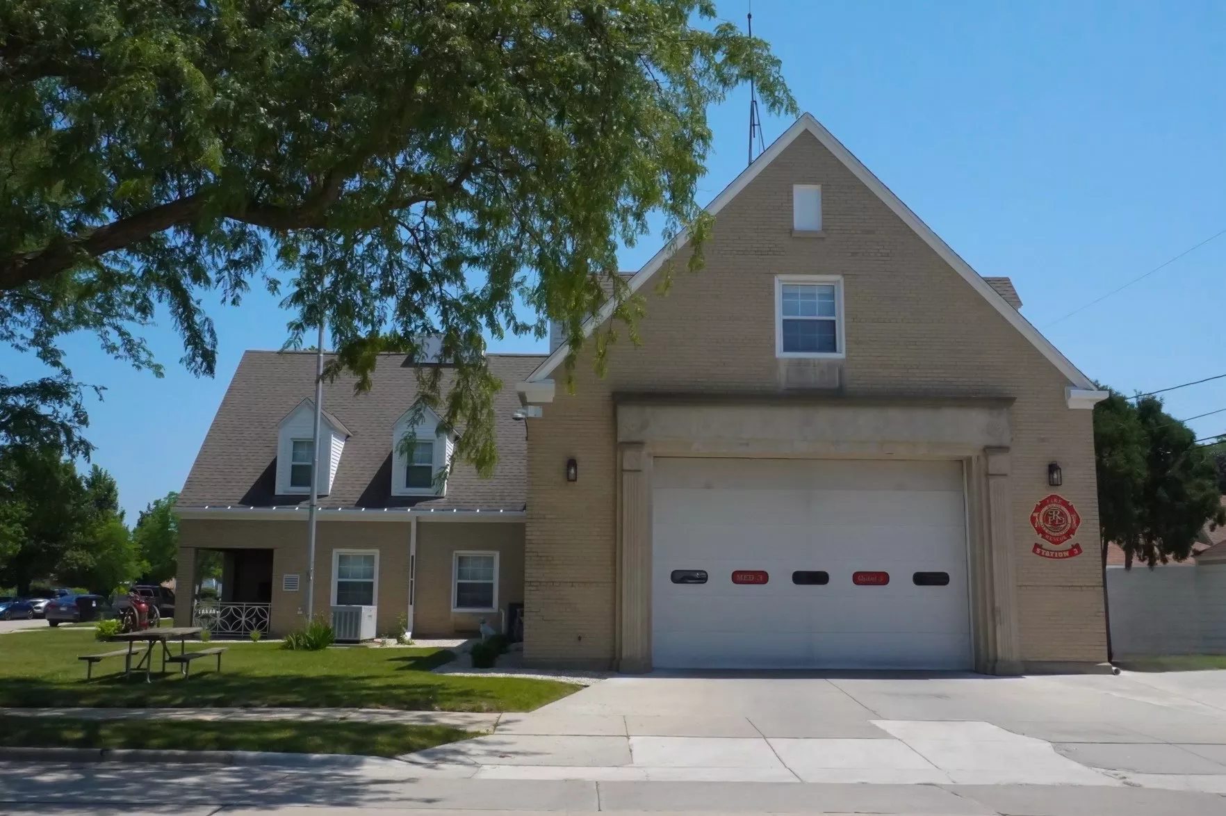 Exterior of a two-story fire station with a large garage door and a peaked roof on a sunny day.