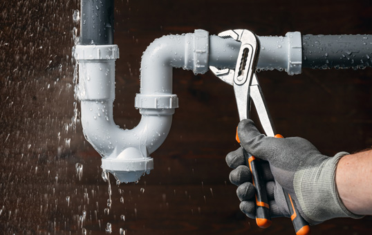 Plumber using adjustable wrench to fix a leaky PVC pipe under a sink. Water droplets visible.