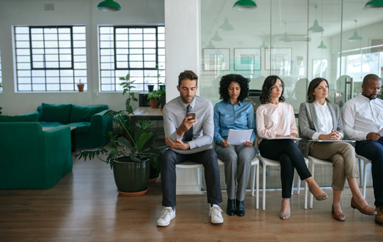 Job applicants sit in a waiting room. One man uses his phone while others hold papers.