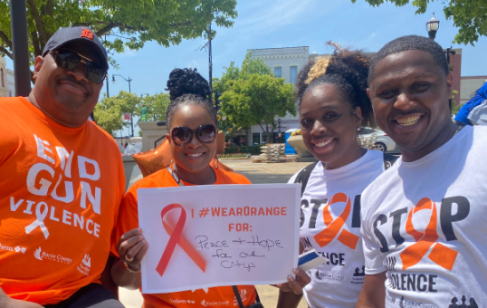 Group of four people wearing orange and white shirts supporting gun violence awareness, holding a sign.