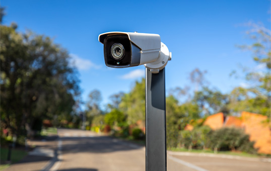 Security camera mounted on a pole, facing forward with a blurred background of trees and a road.