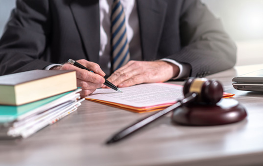 Lawyer reviews legal documents with a pen, gavel, and law books on a desk. Focus on hands and paperwork.