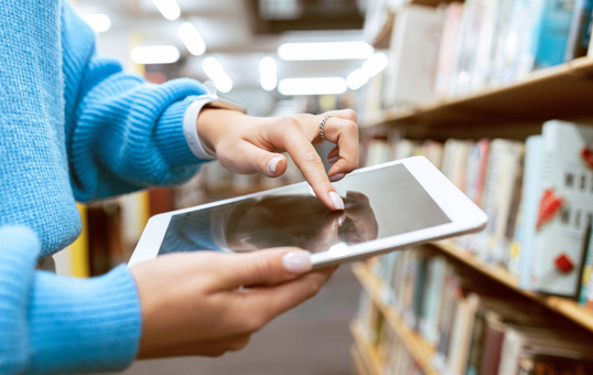Person using a tablet in a library aisle, browsing books on shelves in the background.