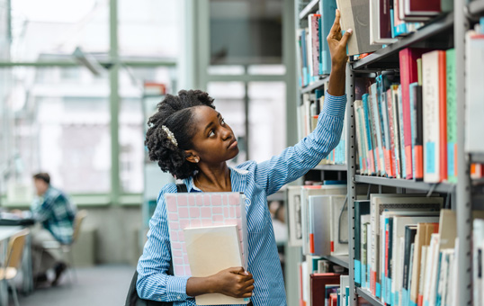 Student reaches for a book on a library shelf, holding notebooks. Another student sits at a table in the background.