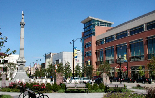 Downtown area with monument, modern buildings, benches, and landscaping on a sunny day.