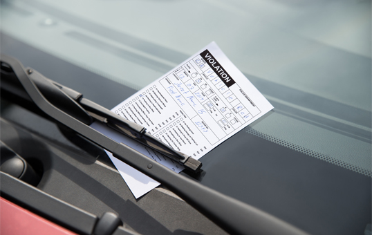 Parking violation ticket under a car's windshield wiper. The ticket is white with black text.