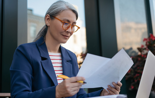 Mature woman with glasses reviews documents outdoors, wearing a blue blazer and striped shirt.