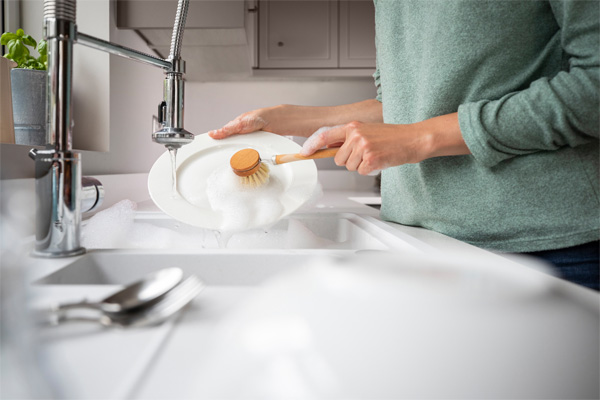 Person washing a white plate with a wooden brush in a white kitchen sink filled with soapy water.
