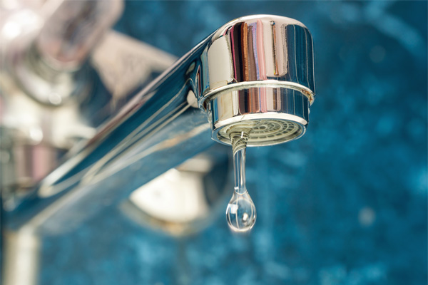 Close-up of a chrome faucet with a single drop of water suspended from the spout against a blue background.