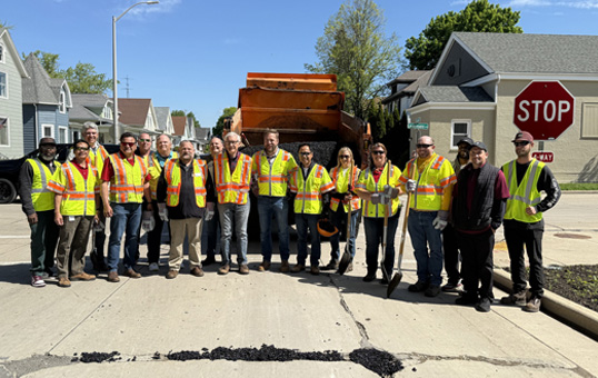 Group of road workers in safety vests stand in front of a dump truck on a residential street.