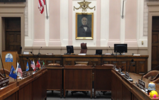 Interior view of a government chamber with flags, desks, and a portrait on the wall.