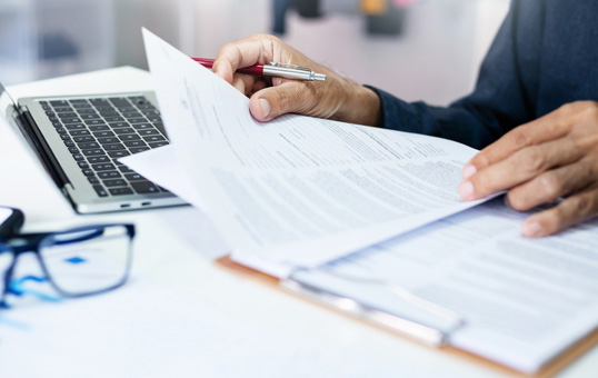 Person reviewing documents with a pen in hand, laptop and glasses on desk. Focus on paperwork.