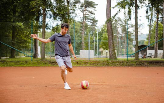 Man in athletic wear kicks a soccer ball on a clay court in a park setting.