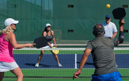Four people playing pickleball on an outdoor court with a green fence. Two yellow balls are in the air.