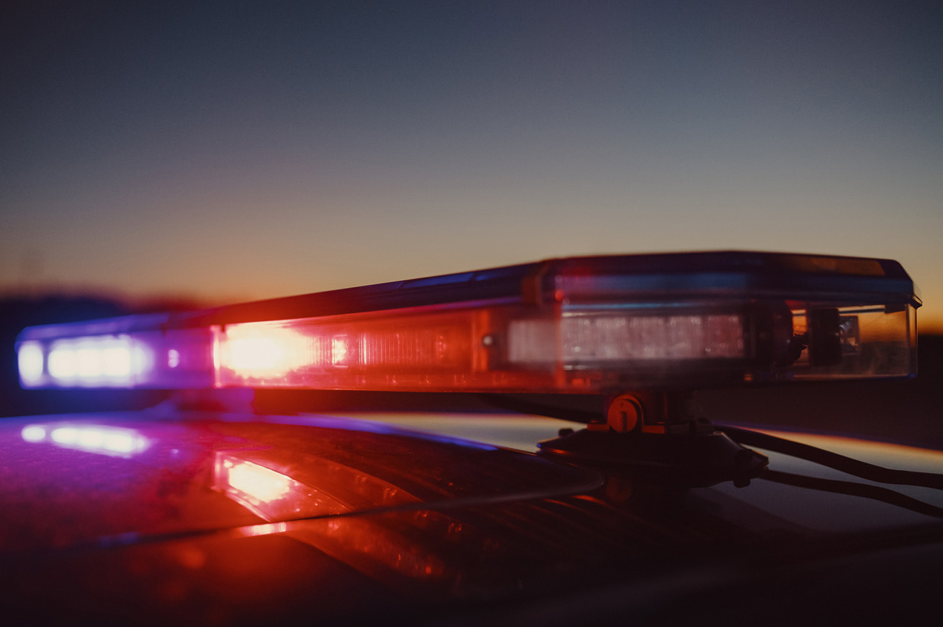 Close-up of police car lights, red and blue, reflecting on the car roof against a dusk sky.
