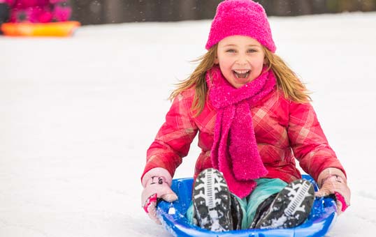 Girl smiling while sledding downhill in the snow, wearing pink hat and scarf.