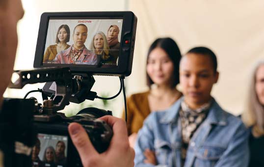 Behind-the-scenes shot of a diverse group of women being filmed with a professional video camera.