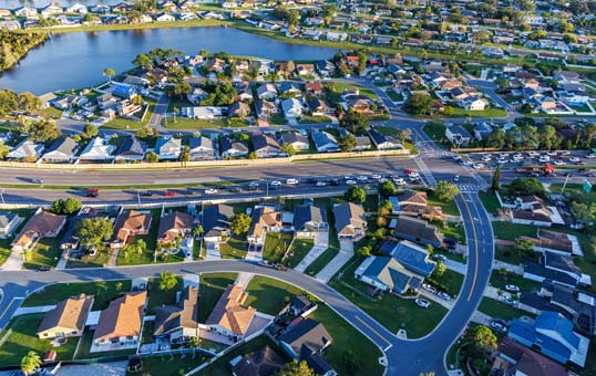 Aerial view of a suburban neighborhood with houses, roads, and a small lake in the background.