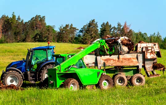 Blue tractor and green loader working together to spread manure on a grassy field. Trees are visible in the background.
