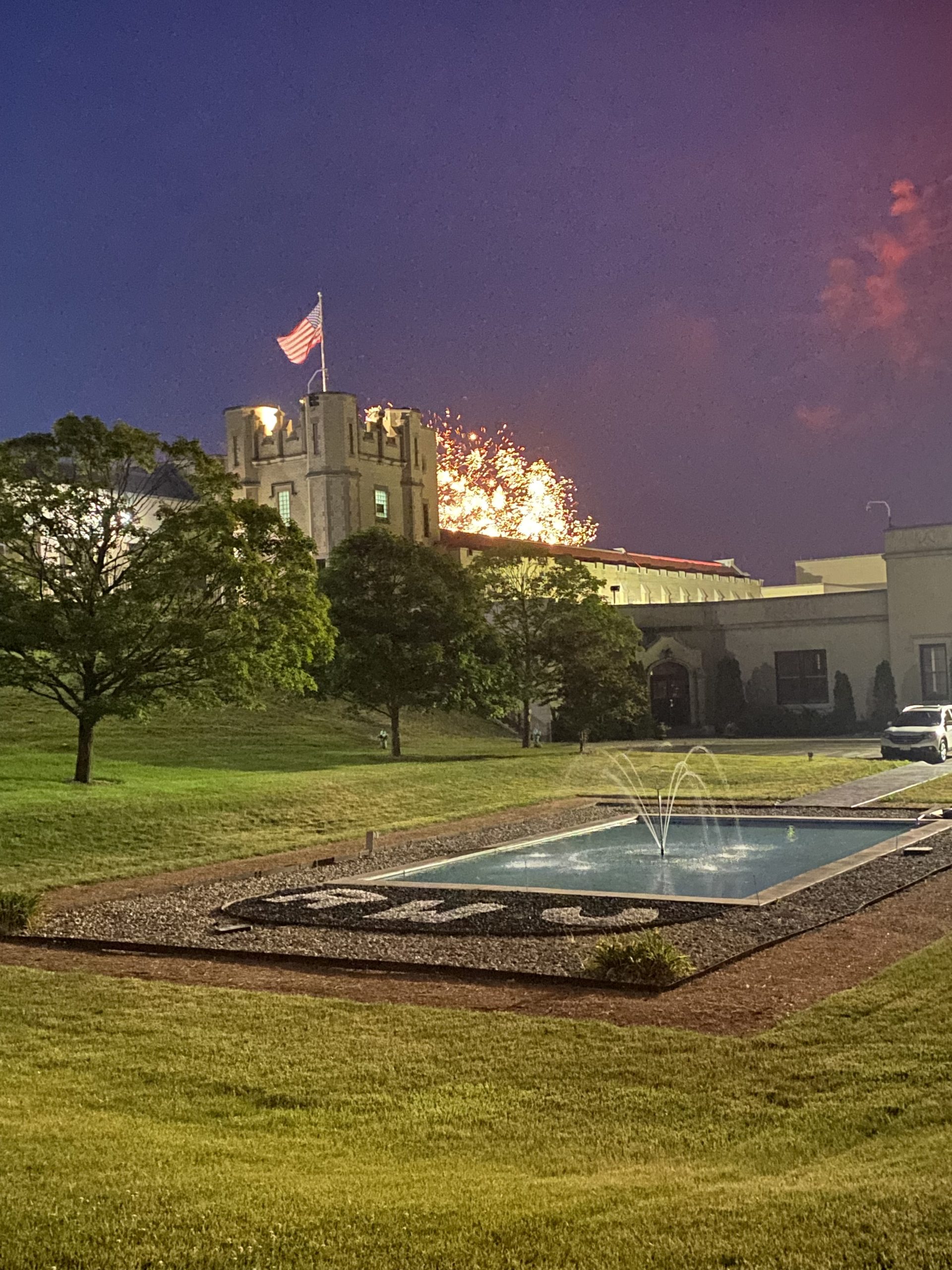 West Point building with fireworks, American flag, fountain, and green lawn at night.