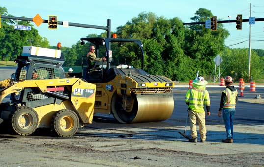 Road construction crew using heavy equipment to repair asphalt at a traffic intersection.