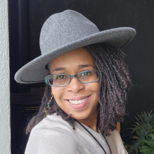 Headshot of a smiling woman with locs wearing a gray hat and glasses.