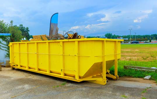 Bright yellow roll-off dumpster filled with debris on a concrete pad, against a cloudy sky.
