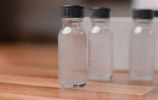 Three clear glass bottles with black caps, standing on a wooden surface, containing a clear liquid.