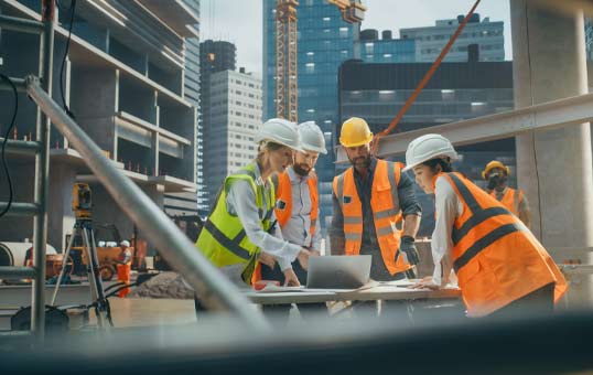 Construction team in hard hats reviewing plans on a laptop at a building site with city skyline in the background.
