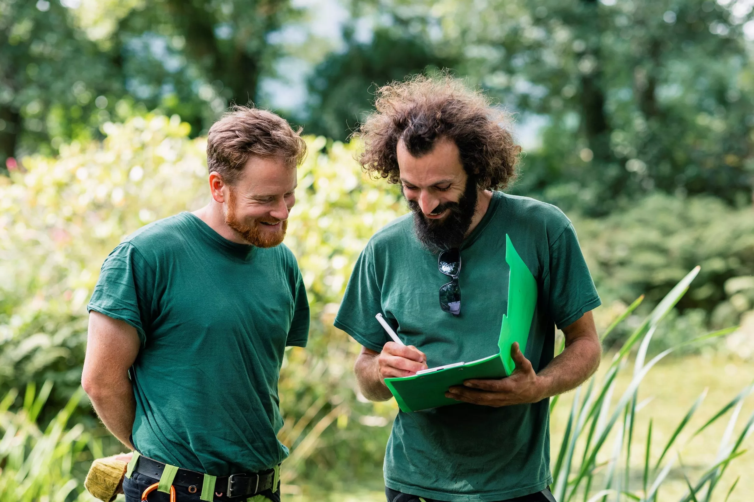 Two groundskeepers in green shirts review notes on a clipboard in a garden setting.