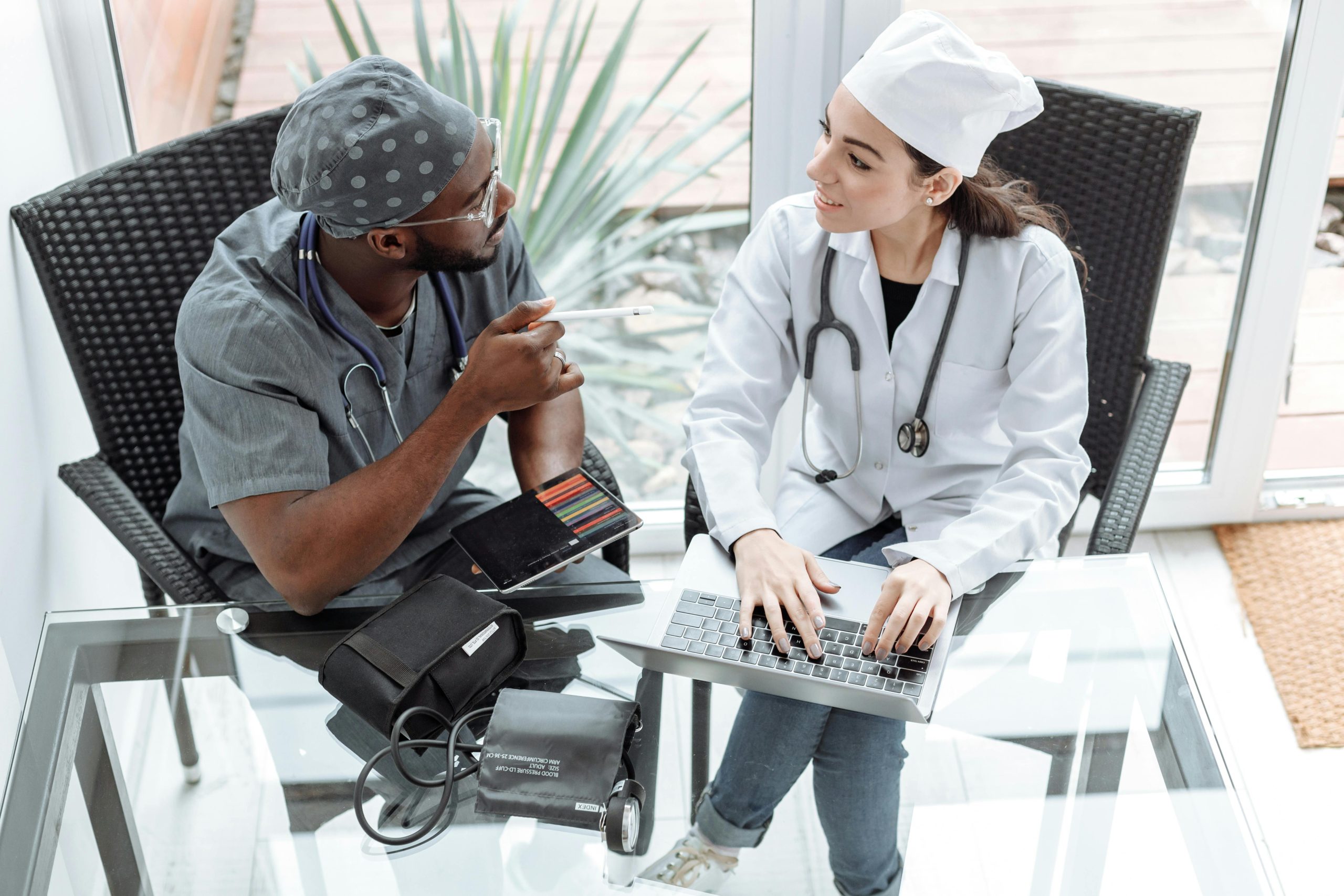 Two healthcare professionals, one using a laptop, discuss patient care at a glass table with medical equipment.