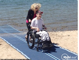Person in wheelchair using Mobi-mat beach access mat with assistance from another person.