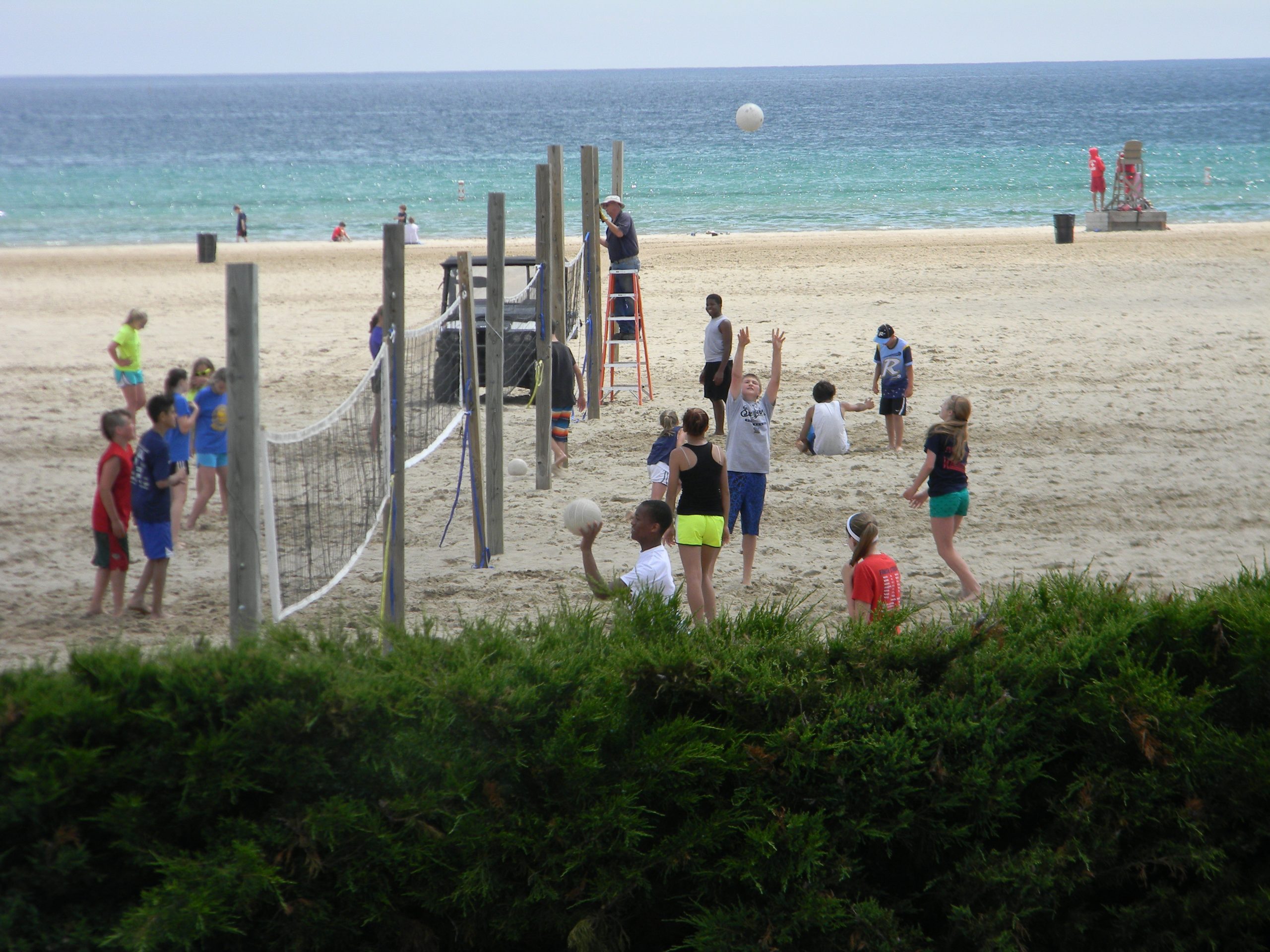 Group of children playing volleyball on a sandy beach with ocean and sky in the background.