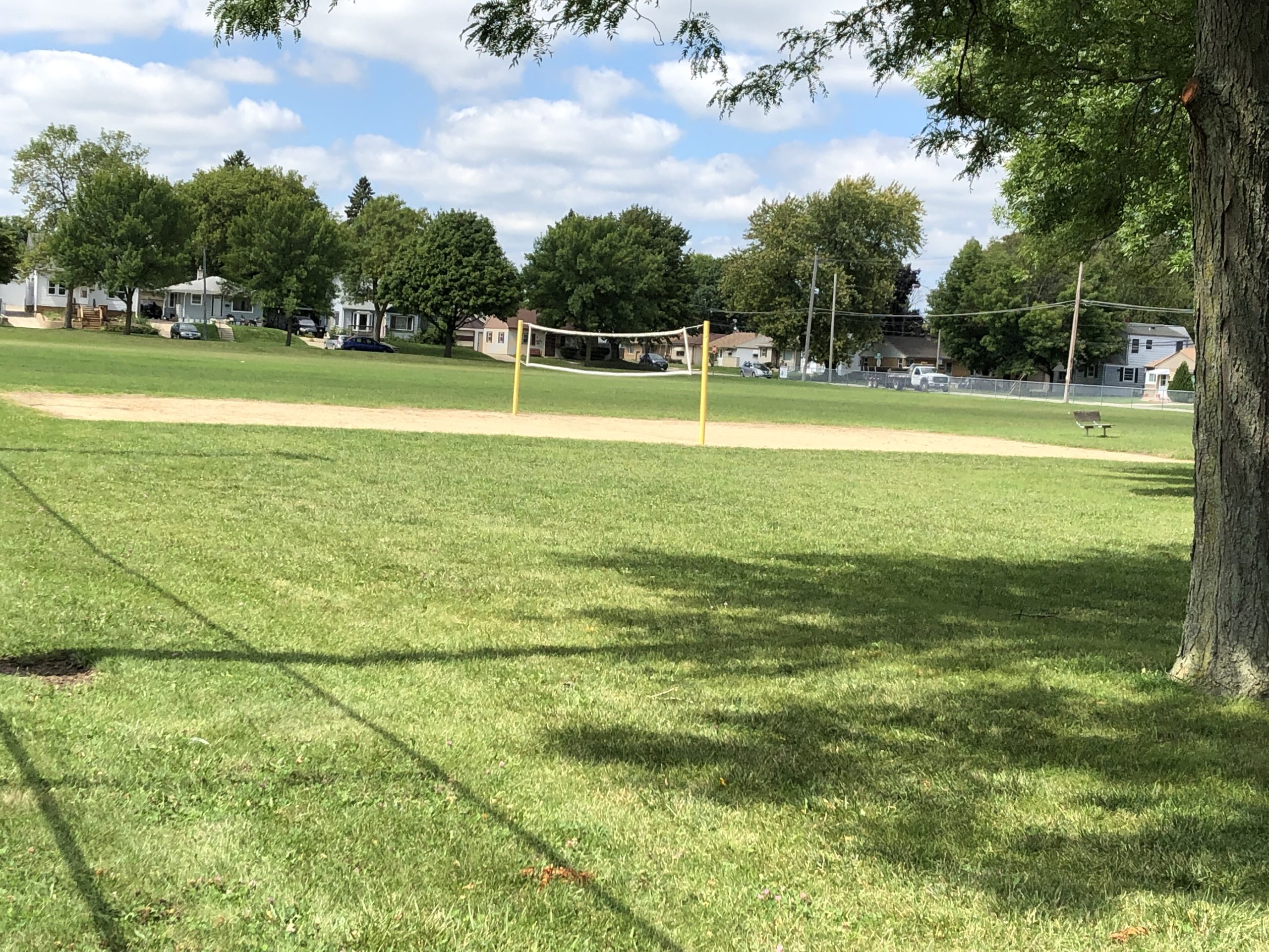Volleyball net stands on a grassy field in a park on a sunny day. Houses and trees are visible in the background.